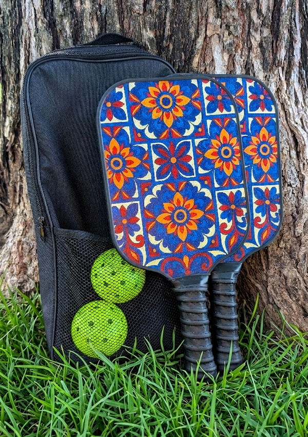 A bright red, blue and yellow floral designed Pickleball paddle set are leaned against a tree with a black carry bag and two pickle balls nicely tucked in place. 